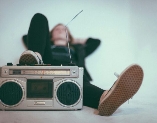 Rádio gravador (boombox) prateado com alto-falantes redondos em primeiro plano, com um pé de tênis estilo lona apoiado em cima. Uma mulher desfocada está deitada e relaxada ao fundo, ouvindo música.