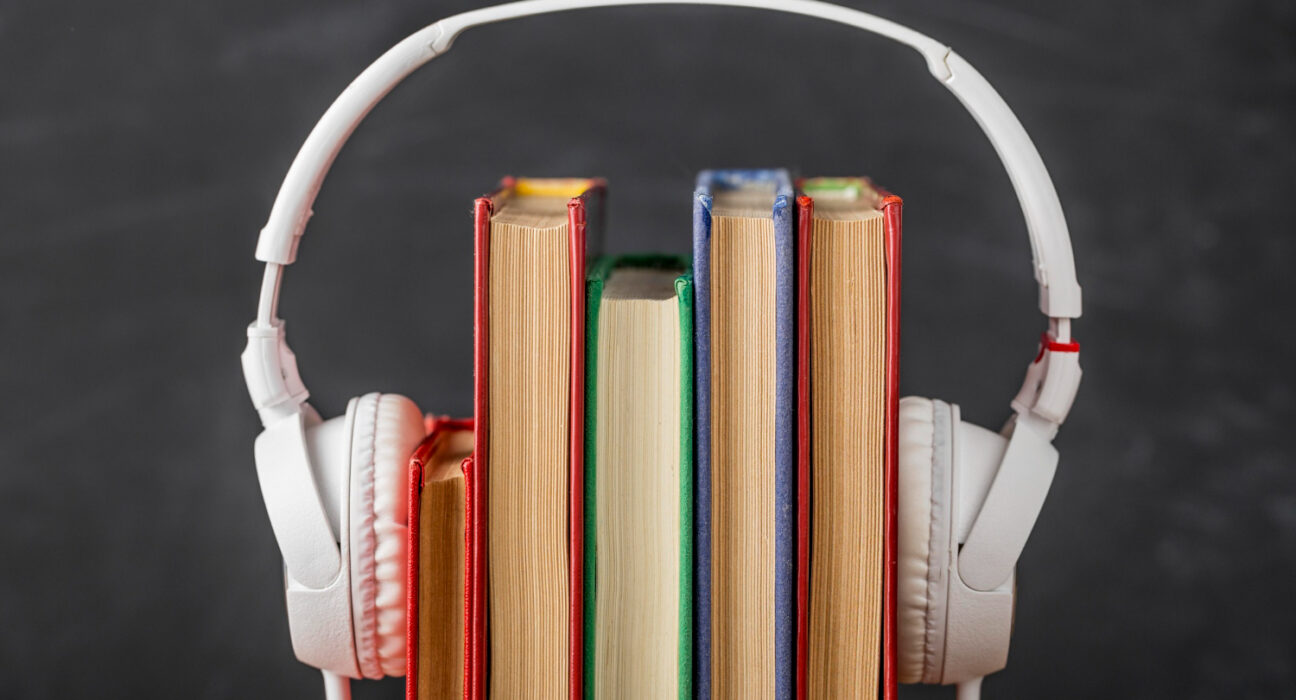 Um par de fones de ouvido brancos sobre uma pilha de cinco livros com lombadas coloridas, em frente a um fundo escuro e desfocado de um quadro-negro