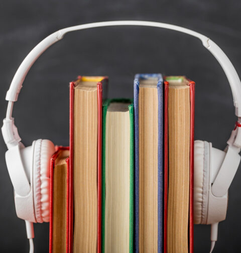 Um par de fones de ouvido brancos sobre uma pilha de cinco livros com lombadas coloridas, em frente a um fundo escuro e desfocado de um quadro-negro