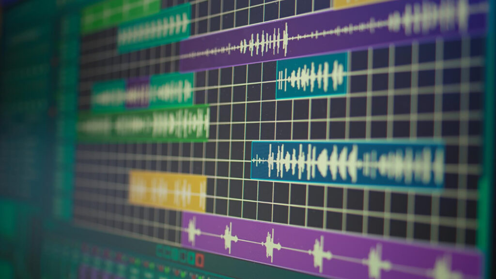 Close-up da tela de um computador exibindo um software de edição de áudio. Várias faixas coloridas em roxo, verde, azul e amarelo mostram formas de onda sonoras sobre uma grade cinza