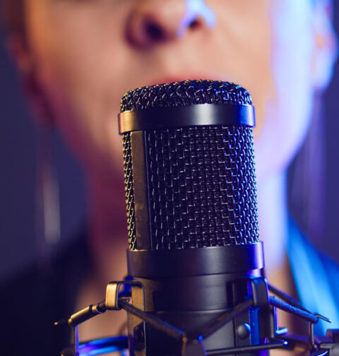 Close-up em um microfone condensador preto de estúdio. O foco está na grade metálica do microfone, enquanto ao fundo, de forma desfocada, aparece o rosto de uma pessoa cantando. A iluminação é dramática, com tons de azul e roxo neon