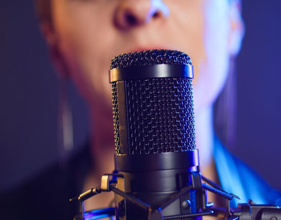 Close-up em um microfone condensador preto de estúdio. O foco está na grade metálica do microfone, enquanto ao fundo, de forma desfocada, aparece o rosto de uma pessoa cantando. A iluminação é dramática, com tons de azul e roxo neon