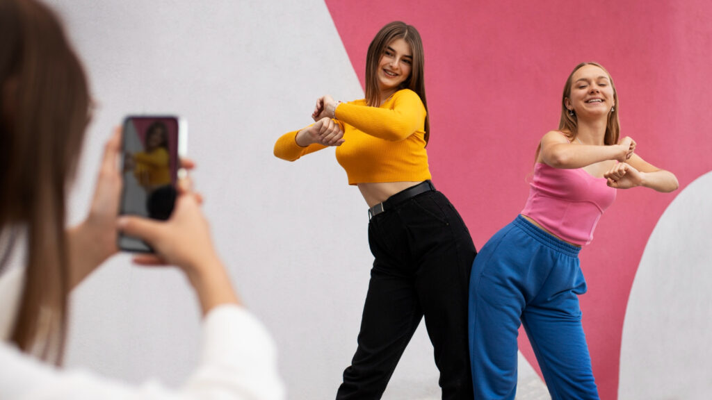 Duas jovens sorridentes fazem uma coreografia de dança em frente a um muro rosa e branco. Em primeiro plano, uma terceira pessoa segura um celular, gravando a performance das duas