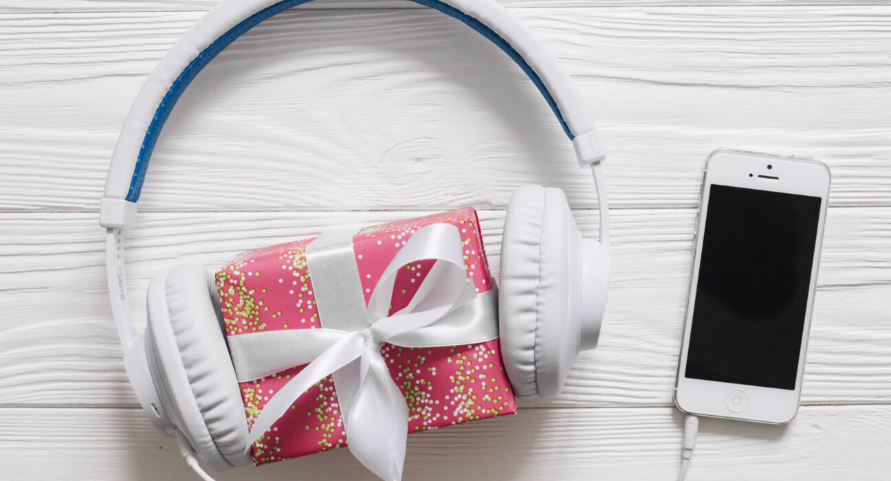 Fones de ouvido brancos envolvendo uma pequena caixa de presente rosa com laço branco, ao lado de um smartphone branco sobre uma superfície de madeira clara