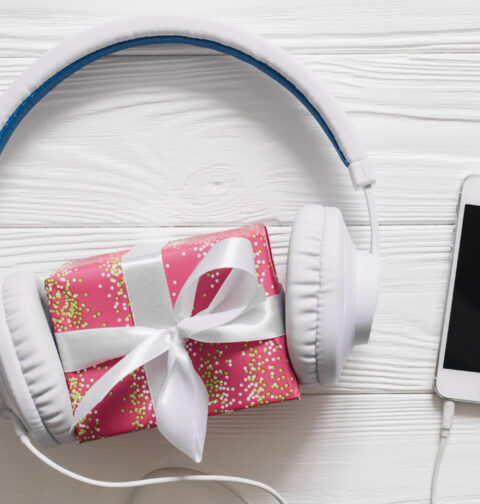 Fones de ouvido brancos envolvendo uma pequena caixa de presente rosa com laço branco, ao lado de um smartphone branco sobre uma superfície de madeira clara