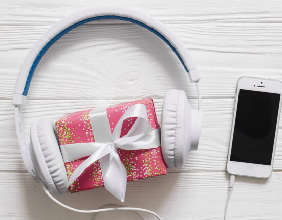 Fones de ouvido brancos envolvendo uma pequena caixa de presente rosa com laço branco, ao lado de um smartphone branco sobre uma superfície de madeira clara