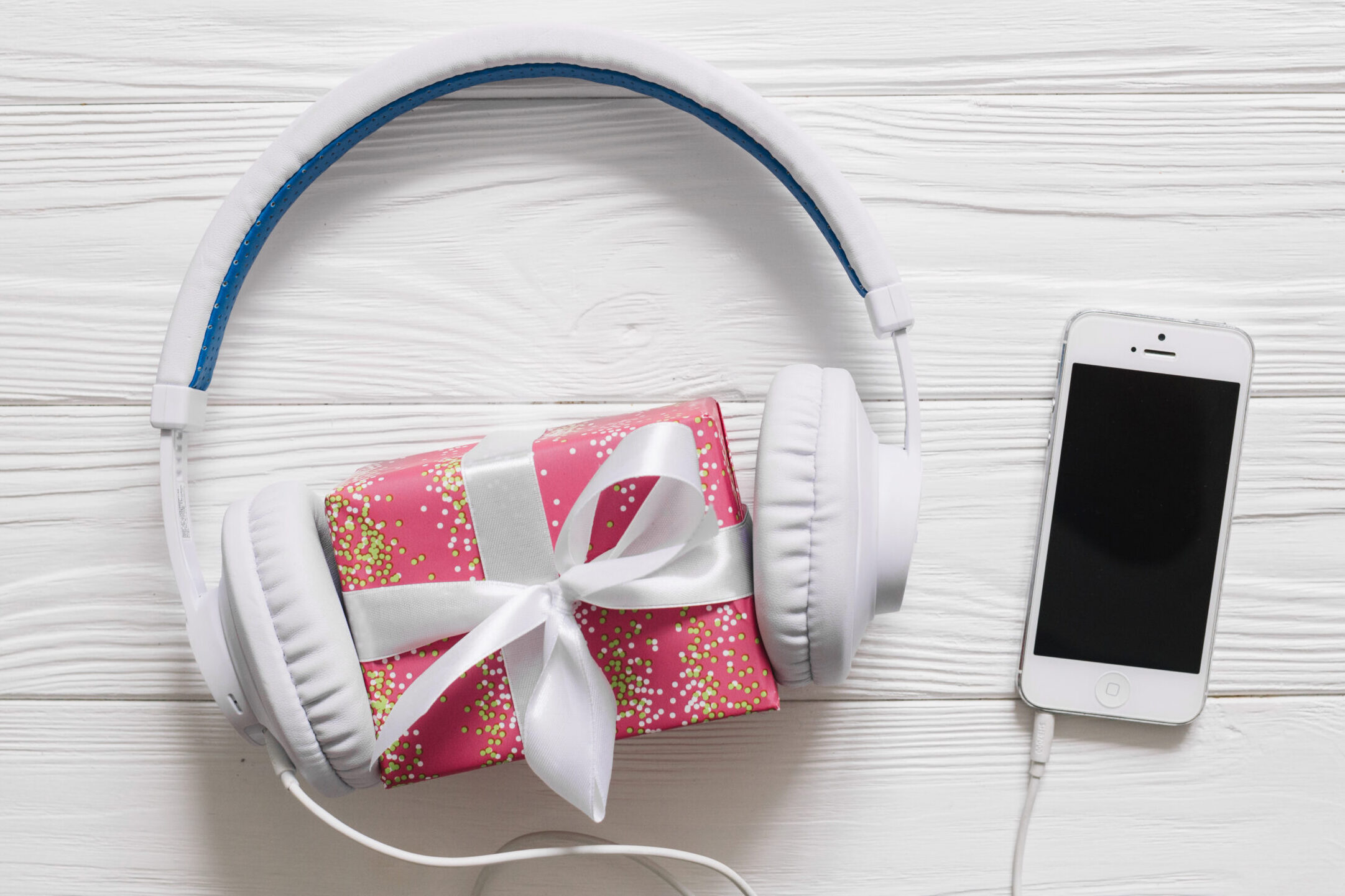 Fones de ouvido brancos envolvendo uma pequena caixa de presente rosa com laço branco, ao lado de um smartphone branco sobre uma superfície de madeira clara