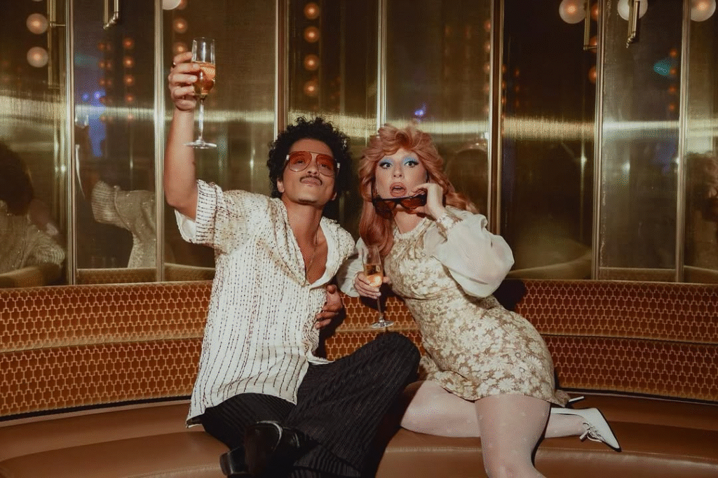 Bruno Mars e Lady Gaga posam sentados em um sofá luxuoso de veludo marrom. Bruno, à esquerda, usa óculos escuros e uma camisa branca brilhante, brindando com uma taça de espumante. Gaga, à direita, usa uma peruca ruiva volumosa, maquiagem azul marcante e um vestido curto floral retrô, também segurando uma taça. O fundo tem painéis dourados e espelhados