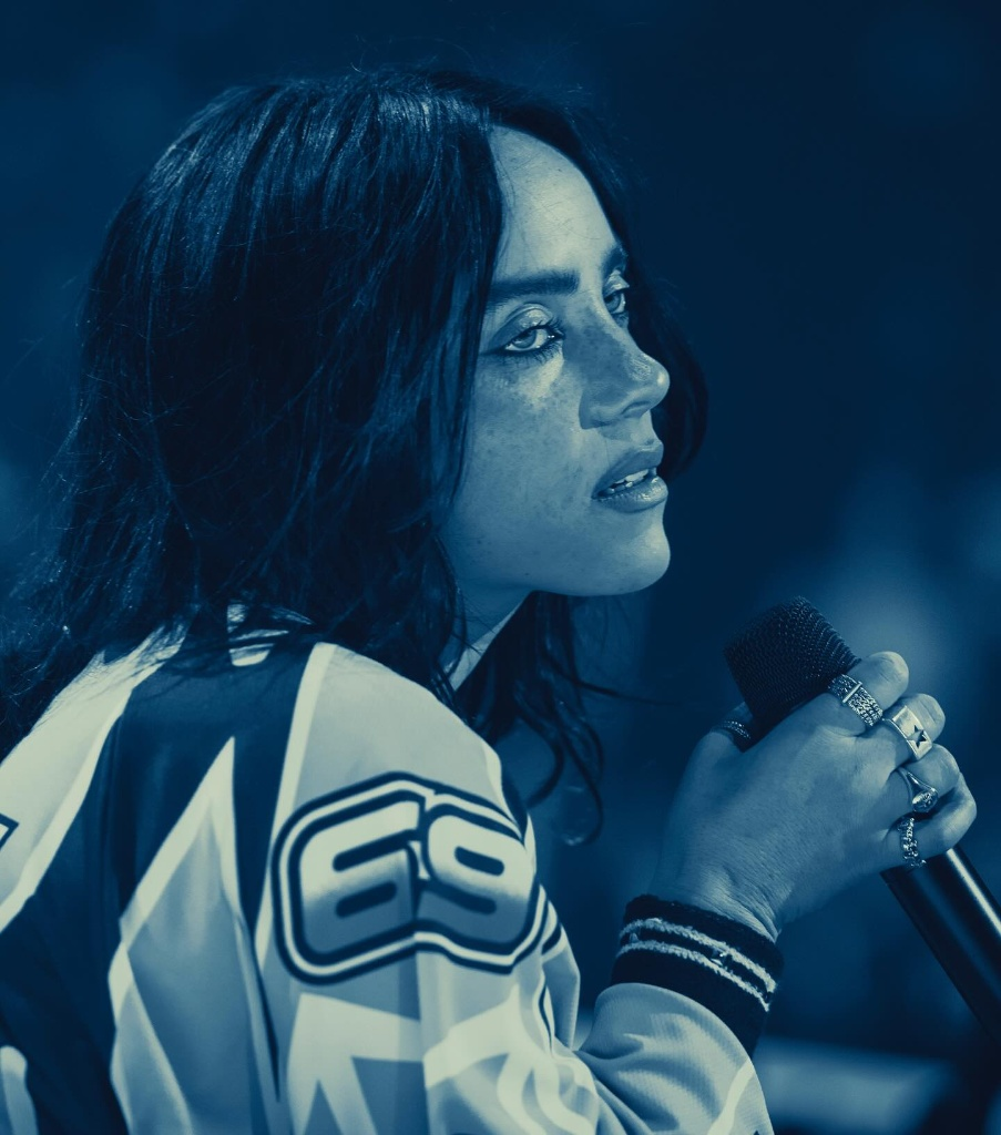 Retrato de perfil da cantora Billie Eilish em tons de azul. Ela tem cabelos escuros, usa uma jaqueta esportiva com o número 69 na manga e segura um microfone próximo ao rosto. Ela olha para o lado com uma expressão pensativa. Seus dedos estão adornados com vários anéis prateados