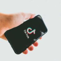 Mão segurando um smartphone com a tela preta exibindo o logotipo colorido do TikTok e o nome da marca em branco. O fundo é neutro e desfocado