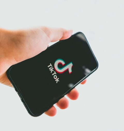 Mão segurando um smartphone com a tela preta exibindo o logotipo colorido do TikTok e o nome da marca em branco. O fundo é neutro e desfocado