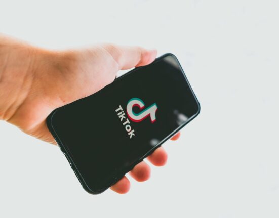 Mão segurando um smartphone com a tela preta exibindo o logotipo colorido do TikTok e o nome da marca em branco. O fundo é neutro e desfocado
